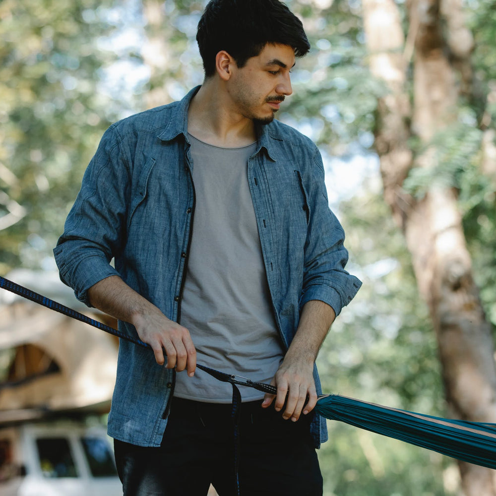 Man adjusting a hammock in a forest setting, wearing a blue shirt and gray t-shirt, surrounded by trees.