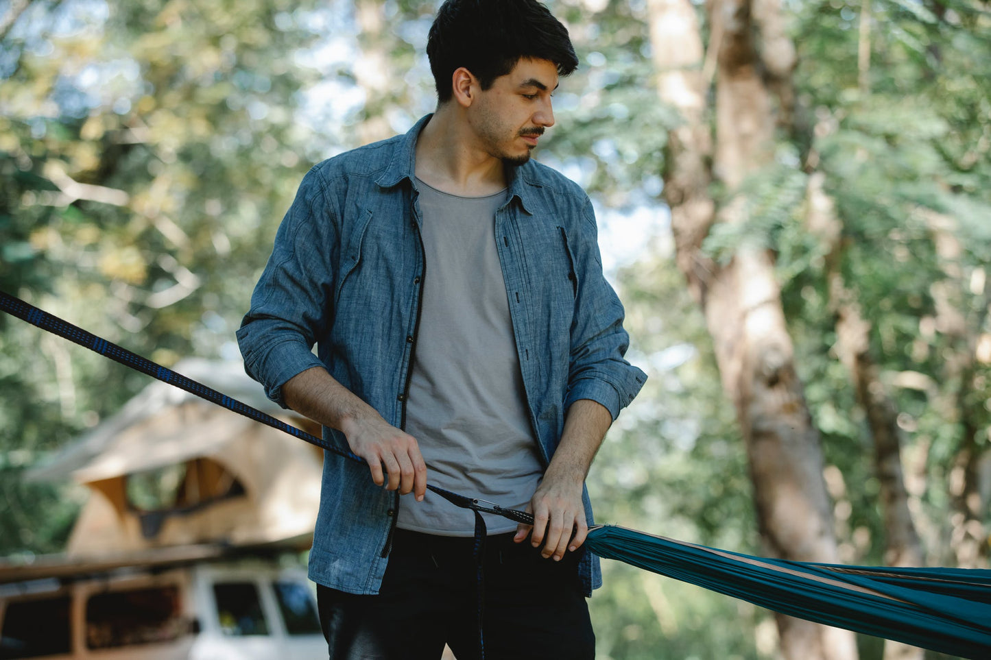 Man adjusting a hammock in a forest setting, wearing a blue shirt and gray t-shirt, surrounded by trees.