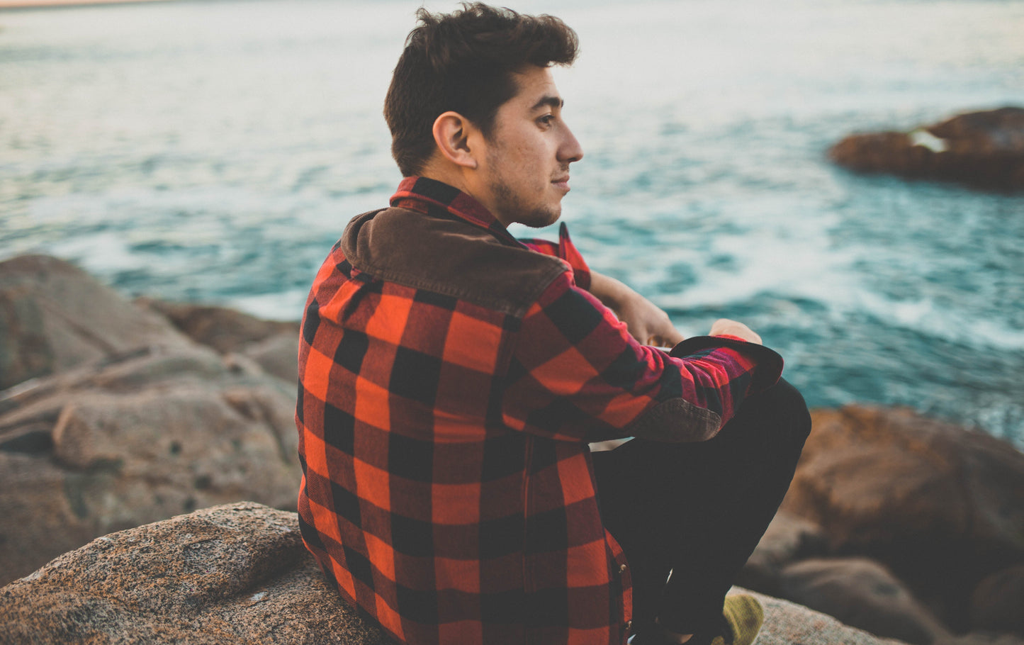 Young man in plaid shirt sitting on rocks by the water, enjoying a peaceful moment outdoors.