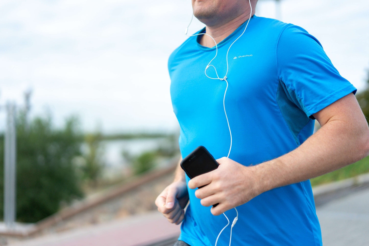 Man running outdoors in a blue athletic shirt, listening to music with earbuds and holding a smartphone.